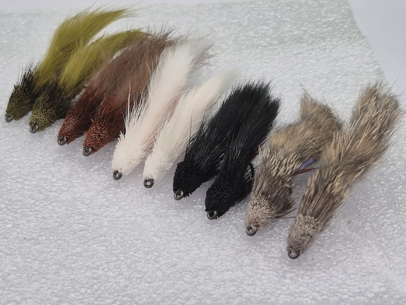 May include: A selection of fly fishing lures in shades of green, brown, white, black, and grey. Each lure features a textured body and a small metal eyelet at the front.