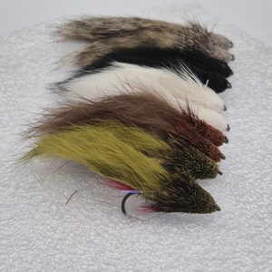 May include: A selection of handmade fishing flies in shades of brown, black, white, and olive green. Each fly is crafted with a hook and designed to resemble insects, attracting fish. The flies are displayed on a white background.
