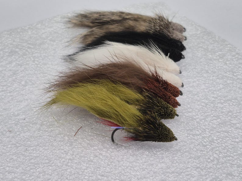 May include: A selection of handmade fishing flies in shades of brown, black, white, and olive green. Each fly is crafted with a hook and designed to resemble insects, attracting fish. The flies are displayed on a white background.