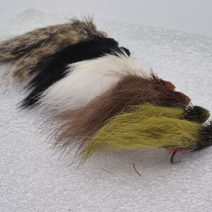 May include: A selection of fishing flies in a row. The flies display a range of colours, including brown, black, white, and olive green. Each fly is equipped with a hook and constructed from diverse materials.