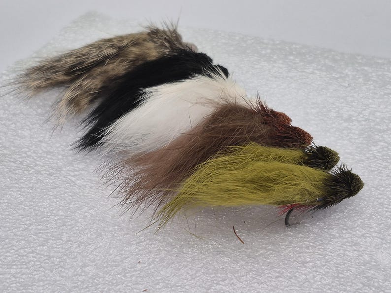 May include: A selection of fishing flies in a row. The flies display a range of colours, including brown, black, white, and olive green. Each fly is equipped with a hook and constructed from diverse materials.