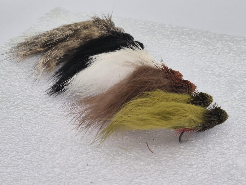 May include: A selection of fishing flies in a row. The flies display a range of colours, including brown, black, white, and olive green. Each fly is equipped with a hook and constructed from diverse materials.