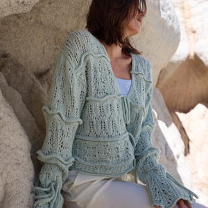Könnte beinhalten: Eine hellsalbeigrüne Langarmstrickjacke mit einem komplizierten Strickmuster, einem V-Ausschnitt und horizontalen Rüschendetails an Oberteil und Ärmeln. Das Kleidungsstück verfügt über einen Bindegürtel an der Vorderseite und ausgestellte Ärmelbündchen.