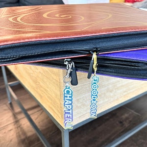 Zipper Pulls to organize Lorcana TCG binders in iridescent color. Available for Lorcana, Chapter 1, Floodborn, Inkands, and Ursula set names and in gold and silver clasps