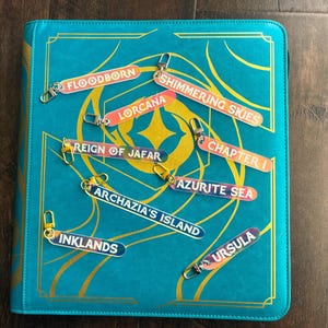 Zipper Pulls for all 8 lorcana sets to organize Lorcana TCG binders in iridescent color. Shown on a Teal Lorcana binder from Silly Bear Emporium