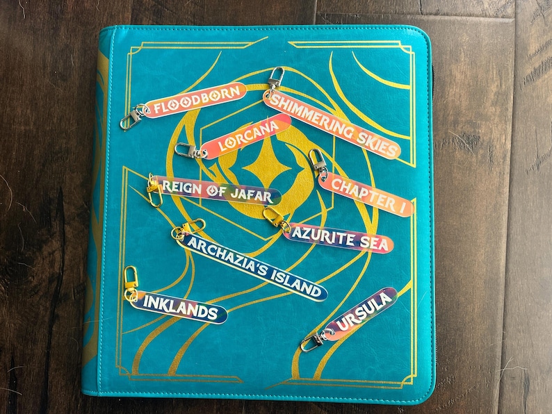 Zipper Pulls for all 8 lorcana sets to organize Lorcana TCG binders in iridescent color. Shown on a Teal Lorcana binder from Silly Bear Emporium