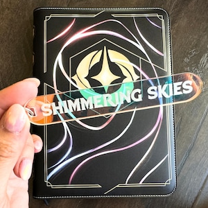 Op de afbeelding: Een zwart lederen gebonden dagboek met een gouden rand en een holografisch ontwerp. Het dagboek heeft een bladwijzer met de tekst "Shimmering Skies".
