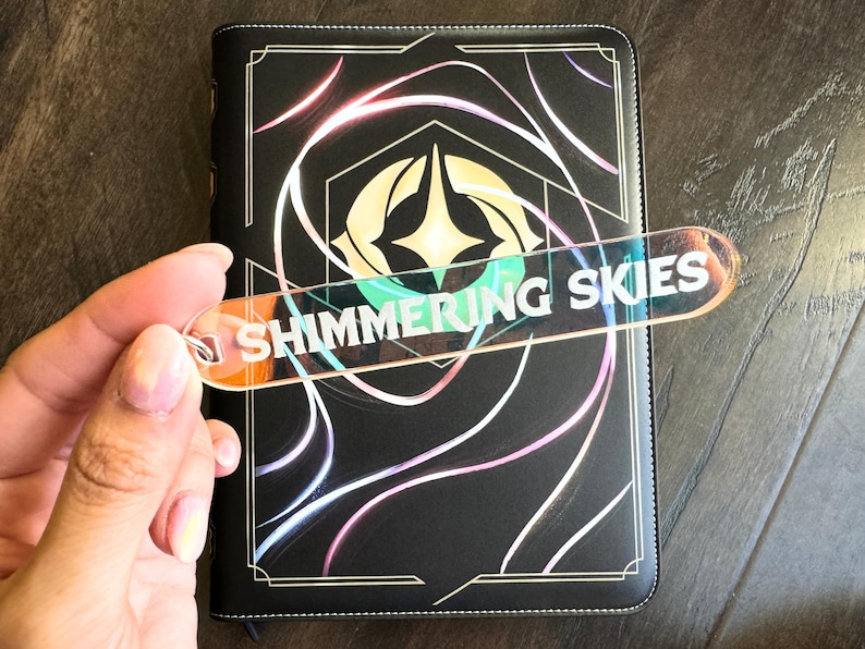 Op de afbeelding: Een zwart lederen gebonden dagboek met een gouden rand en een holografisch ontwerp. Het dagboek heeft een bladwijzer met de tekst "Shimmering Skies".