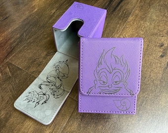 May include: Purple card case with a flap closure, featuring a black line drawing of a cartoon character. The interior has a gray lining with a matching illustration. A matching box is also visible.