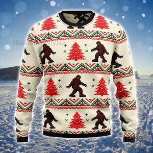May include: A cream-coloured long-sleeve sweater featuring a repeating pattern of brown Bigfoot silhouettes and red Christmas trees. The design includes horizontal bands of green and red geometric shapes, with a black crew neck and cuffs.