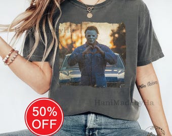 May include: Dark gray t-shirt featuring a graphic of a character from a horror movie making a heart shape with his hands. The image is set against a blurred background. The text "HaniMadebyHa" is printed on the shirt. A red circle with "50% OFF" is in the lower left.