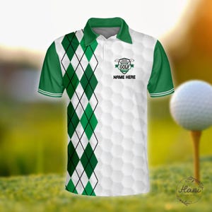 Puede incluir: Polo de golf blanco y verde con un patrón de diamantes en el lado izquierdo. La camisa tiene un logotipo con temática de golf y el texto "NAME HERE". Una pelota de golf en un tee está en el fondo.