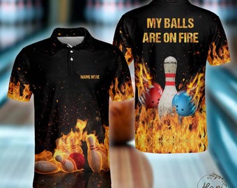 May include: Black polo shirt with a bowling theme. The shirt features a design of bowling pins, red and blue bowling balls, and flames. The text "MY BALLS ARE ON FIRE" is printed on the back. The front of the shirt has the text "NAME HERE".