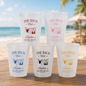 May include: Five frosted plastic cups featuring the text THE BACH CLUB EST. 2026, a graphic of bikinis on a clothesline, and the text Sophia's BACHELORETTE NEW YORK, NY. Each cup displays the design in a different color: red, pink, blue, black, and gold.
