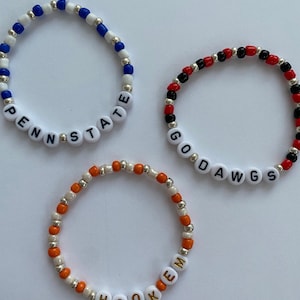 Peut inclure: Trois bracelets de perles avec des perles de lettres blanches épelant "PENN STATE", "GO DAWGS" et "HOOKEM". Les bracelets présentent des perles bleues, rouges, oranges et blanches, avec des perles d'accent argentées. Ces bracelets sont parfaits pour montrer l'esprit d'équipe.