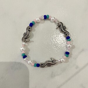 May include: A beaded bracelet featuring three silver seahorse charms. The bracelet is strung with white, pink, teal, and navy blue beads. The beads are a mix of small round beads and larger faux pearls.