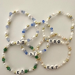 May include: A collection of beaded bracelets featuring white, gold, blue, and green beads. Each bracelet has a different Greek letter combination spelled out in black letters on white beads. The bracelets are arranged in a circular pattern.