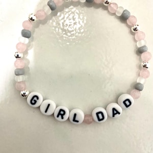 May include: A beaded bracelet with the words "GIRL DAD" spelled out in white letter beads. The bracelet features pink, gray, and clear beads, along with silver-colored accent beads. The bracelet is designed for everyday wear.