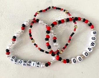 Könnte beinhalten: Drei Armbänder mit Perlen in Rot, Schwarz, Weiß und Gold. Ein Armband buchstabiert "GEORGIA" und ein anderes "GODAWGS" mit weißen Buchstabenperlen. Die Armbänder liegen auf einer weißen Oberfläche.