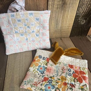 May include: Two quilted fabric bags with different patterns. One bag is white with pink and blue butterflies and the other is white with a floral pattern in shades of pink, blue, yellow, and orange. The floral bag has a brown velvet ribbon.