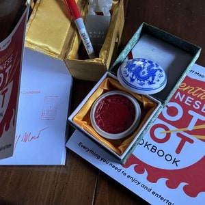 Könnte beinhalten: Zwei Kochbücher mit dem Titel "Essential Chinese Hot Pot Cookbook" auf dem Cover. Das Cover ist rot mit einem weißen Umriss eines Hot Pots. Die Kochbücher sind auf der ersten Seite aufgeschlagen. Ein rotes Stempelkissen befindet sich in einer kleinen Holzkiste mit einem blauen und weißen Porzellandeckel. Der Deckel hat ein Drachenmotiv. Der Text "Essential Chinese Hot Pot Cookbook" ist auf dem Cover des Buches sichtbar.