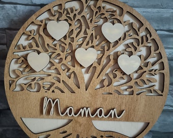 Puede incluir: Un árbol genealógico circular de madera con un diseño cortado con láser. El árbol presenta cinco recortes en forma de corazón y la palabra "Maman" en cursiva. Se apoya en un caballete de madera, mostrando la veta natural de la madera.