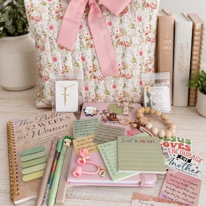 May include: A quilted fabric pouch with a large pink bow, containing a spiral-bound book titled The Bible in 52 Weeks, a gold cross necklace, a wooden bead keychain, pens, sticky notes, and various cards with religious text and illustrations.
