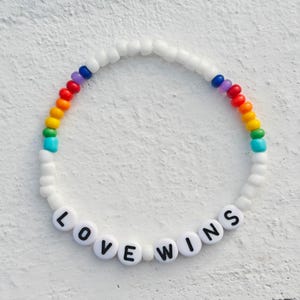 Op de afbeelding: Een witte kralen armband met een regenboog van gekleurde kralen en de woorden "LOVE WINS" gespeld in zwarte letters op witte kralen.