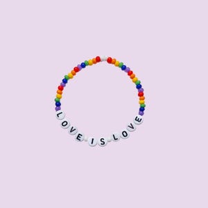 Peut inclure: Un bracelet de perles aux couleurs de l'arc-en-ciel avec les mots "LOVE IS LOVE" épelés en perles de lettres blanches. Le bracelet est posé sur un fond violet clair.