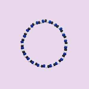 Puede incluir: Una pulsera de cuentas con un patrón de cuentas azules, blancas y negras. La pulsera es circular y se encuentra sobre un fondo lila claro. Las cuentas son pequeñas y están espaciadas uniformemente.