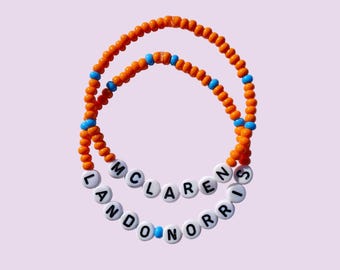 Puede incluir: Dos pulseras de cuentas. La pulsera superior es naranja con detalles azules. La pulsera inferior es blanca con letras negras que deletrean "LANDON NORRI". La pulsera superior tiene las letras "MCLARENS". El fondo es de color lila claro.
