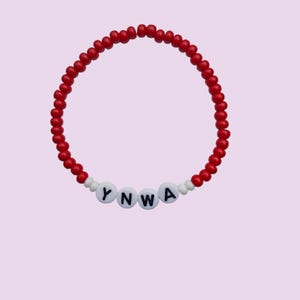 May include: Red beaded bracelet featuring the letters "YNWA". The bracelet is made of small, round red beads and white letter beads. The background is a pale purple colour.