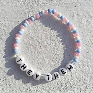 Op de afbeelding: Een kralenarmband met afwisselende blauwe, roze en witte kralen. De armband spelt "THEY THEM" in witte kralen met zwarte letters.
