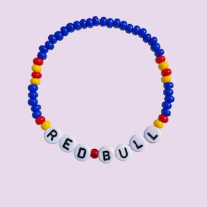 May include: A beaded bracelet with the word "REDBULL" spelled out in white letter beads. The bracelet features blue, red, and yellow beads. The bracelet is on a light purple background.