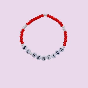May include: A bracelet made of red and white beads. The bracelet features the letters "SL BENFICA" in white beads. The background is a pale pink colour.