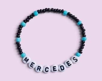 May include: A circular beaded bracelet featuring small black and turquoise beads arranged in a repeating pattern. The lower section includes white spherical beads with the black printed letters spelling out the name MERCEDES.