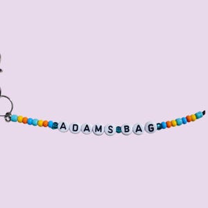 以下が含まれることがあります： 「ADAMS BAG」の文字が黒い文字で白いビーズに書かれたビーズキーホルダー。ビーズは青、オレンジ、黄色のビーズで繋がれています。シルバーカラーの留め金が付いています。