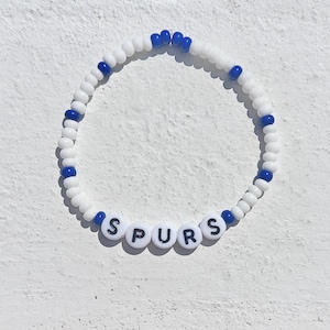 Op de afbeelding: Wit en blauw kralen armband met het woord "SPURS" gespeld in zwarte letters.