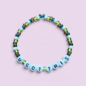 May include: A circular beaded bracelet featuring a repeating pattern of translucent teal, brown, and neon yellow beads. The bracelet includes white spherical beads with black printed letters spelling out the phrase U GOT THIS.