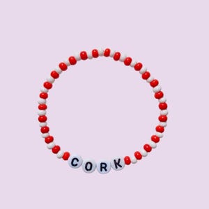 May include: A beaded bracelet with red and white beads. The word "CORK" is spelled out in white letter beads. The bracelet is on a light purple background.