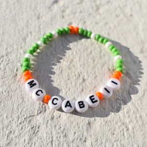 Könnte beinhalten: Ein weißes Perlenarmband mit dem Namen "MCAIBE" in schwarzen Buchstaben. Die Perlen wechseln sich in Grün, Weiß und Orange ab.