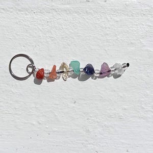 Puede incluir: Un llavero con una serie de pequeñas piedras preciosas de colores en un arreglo de arcoíris. Las piedras son rojas, naranjas, amarillas, verdes, azules, moradas y transparentes. Están ensartadas en un alambre negro y unidas a un anillo de metal plateado.