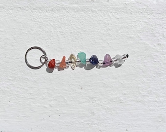 May include: A keychain featuring a series of small, colorful gemstones in a rainbow arrangement. The stones are red, orange, yellow, green, blue, purple, and clear. The stones are strung together on a black wire and attached to a silver metal ring.