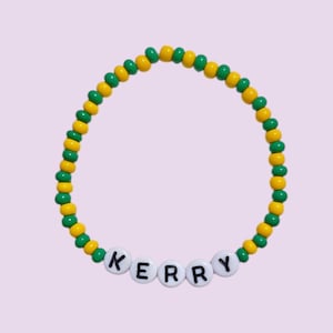 Könnte beinhalten: Ein Armband mit abwechselnd grünen und gelben Perlen. Das Armband schreibt den Namen "KERRY" mit weißen Buchstabenperlen. Das Armband wird auf einem hellvioletten Hintergrund präsentiert.