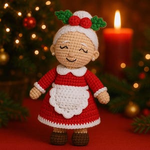 May include: A crocheted Mrs. Claus doll with a red dress, white apron, and a white cap adorned with holly berries. The doll has a smiling face, pearl earrings, and brown shoes. The background includes a Christmas tree and a lit candle.