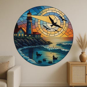 Op de afbeelding: Een ronde wandkunst met een kustscène met een vuurtoren, golven en vliegende vogels. Het kunstwerk heeft een glas-in-loodstijl met levendige kleuren, waaronder blauw, oranje en geel. Twee eenden zwemmen in het water.