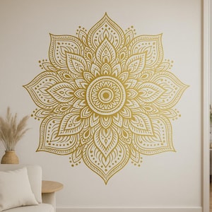 Op de afbeelding: Een grote, goudkleurige mandala muursticker met ingewikkelde bloempatronen. Het symmetrische ontwerp heeft bloemblaadjes en gestippelde accenten. De sticker is op een neutrale muur aangebracht, wat een opvallend contrast creëert.
