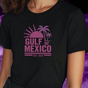 Könnte beinhalten: Schwarzes T-Shirt mit einem lila Aufdruck von Sonne, Palmen und dem Text "GULF OF MEXICO" mit "EST. 1550" darunter. Das T-Shirt ist aus einem weichen Material.