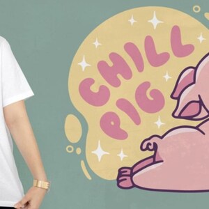 May include: White t-shirt featuring a cartoon pig in a relaxed pose, with the words "CHILL PIG" in pink bubble letters. The pig is light pink, and the background is a yellow circle with white star accents.