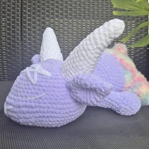 May include: A crocheted stuffed unicorn toy with a purple body, white horn, and a rainbow tail. The unicorn is lying on its side with its head resting on the ground.